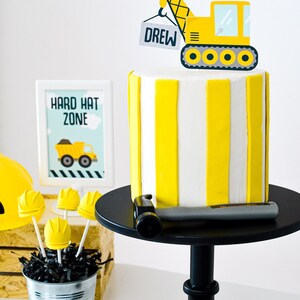 May include: A yellow and white striped cake with a yellow construction crane topper that says "Drew" on it. The cake is on a black cake stand with a hammer on the stand. There is a sign that says "Hard Hat Zone" and a bucket of yellow hard hat cake pops.