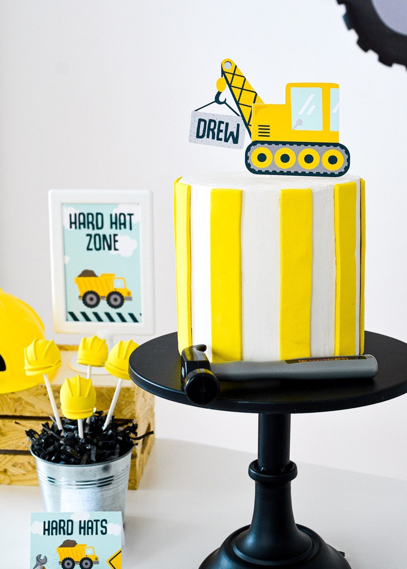 May include: A yellow and white striped cake with a yellow construction crane topper that says "Drew" on it. The cake is on a black cake stand with a hammer on the stand. There is a sign that says "Hard Hat Zone" and a bucket of yellow hard hat cake pops.