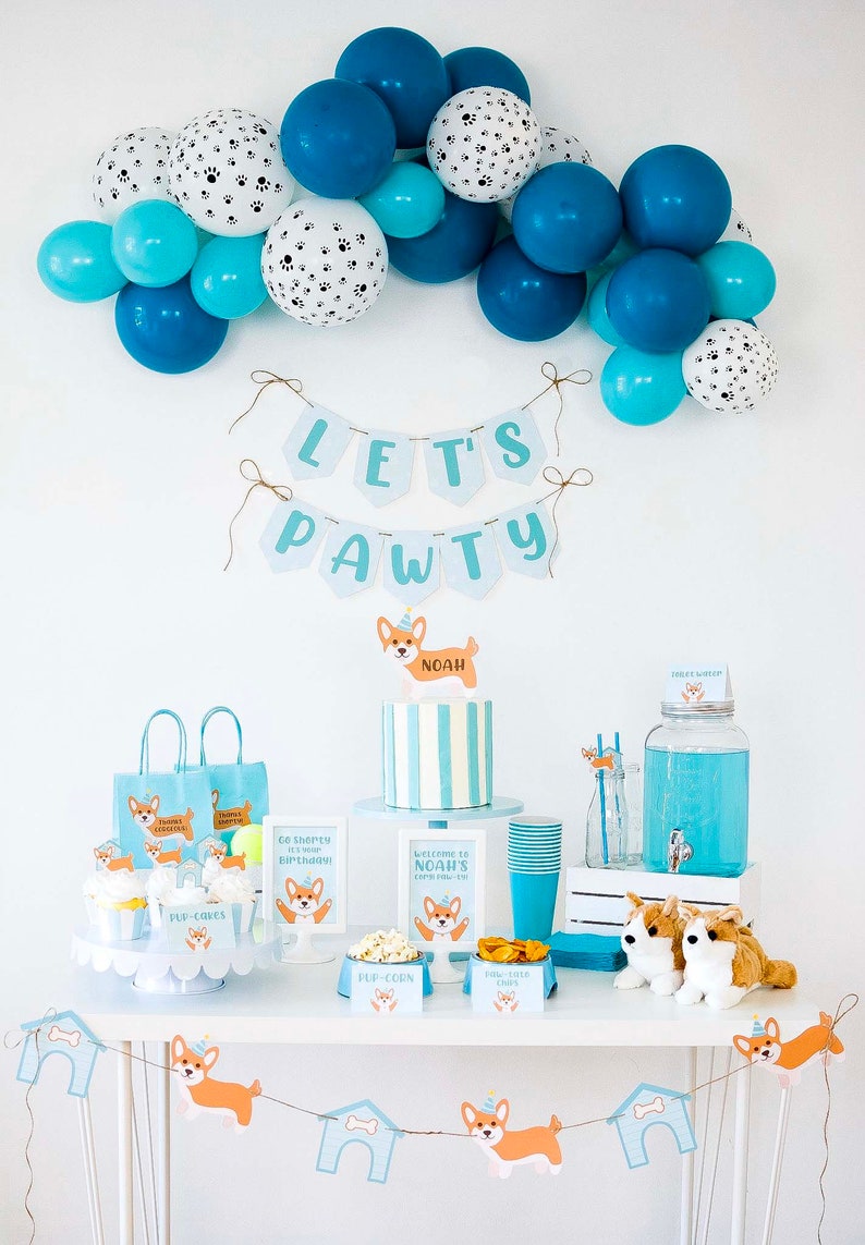 May include: A party setup with a 'Let's Pawty' banner, balloons, and corgi-themed decorations. Includes a cake, cupcakes, gift bags, and signs. The color scheme is blue, white, and orange. The text on the banner reads 'Let's Pawty'.