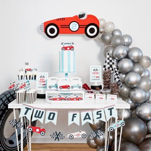 May include: A red and white race car themed party table setting with a cake, cupcakes, and a banner that says "Two Fast". The table is decorated with silver balloons and a checkered flag.