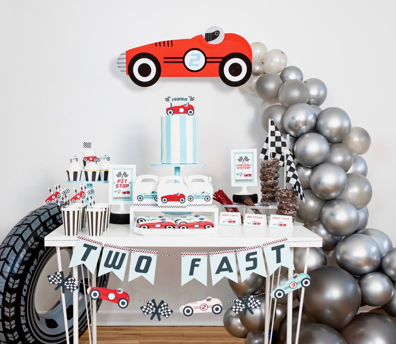 May include: A red and white race car themed party table setting with a cake, cupcakes, and a banner that says "Two Fast". The table is decorated with silver balloons and a checkered flag.