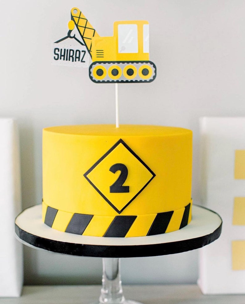 May include: A yellow and black construction themed birthday cake with a number 2 on a yellow diamond sign. The cake is decorated with a yellow construction vehicle topper that says "SHIRAZ" on the side.