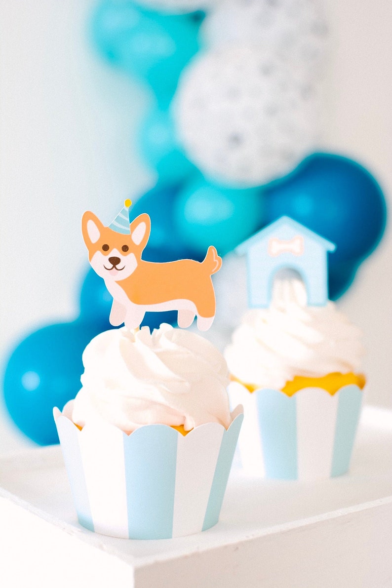May include: Two cupcakes decorated with a dog wearing a party hat and a dog house. The cupcakes are in blue and white striped paper liners.