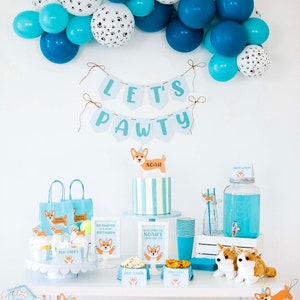 May include: A birthday party table setting with a blue and white color scheme. The table is decorated with a banner that says "Let's Pawty", a cake with a dog topper, and various snacks and drinks. The table is also decorated with a garland of blue and white balloons.