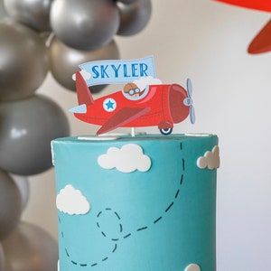 Puede incluir: Una tarta de cumplea&ntilde;os con una base de glaseado azul, decorada con nubes blancas y una trayectoria de vuelo punteada. Un adorno de tarta de avi&oacute;n rojo con el nombre "Skyler" en una pancarta se encuentra encima de la tarta. Globos grises en el fondo.