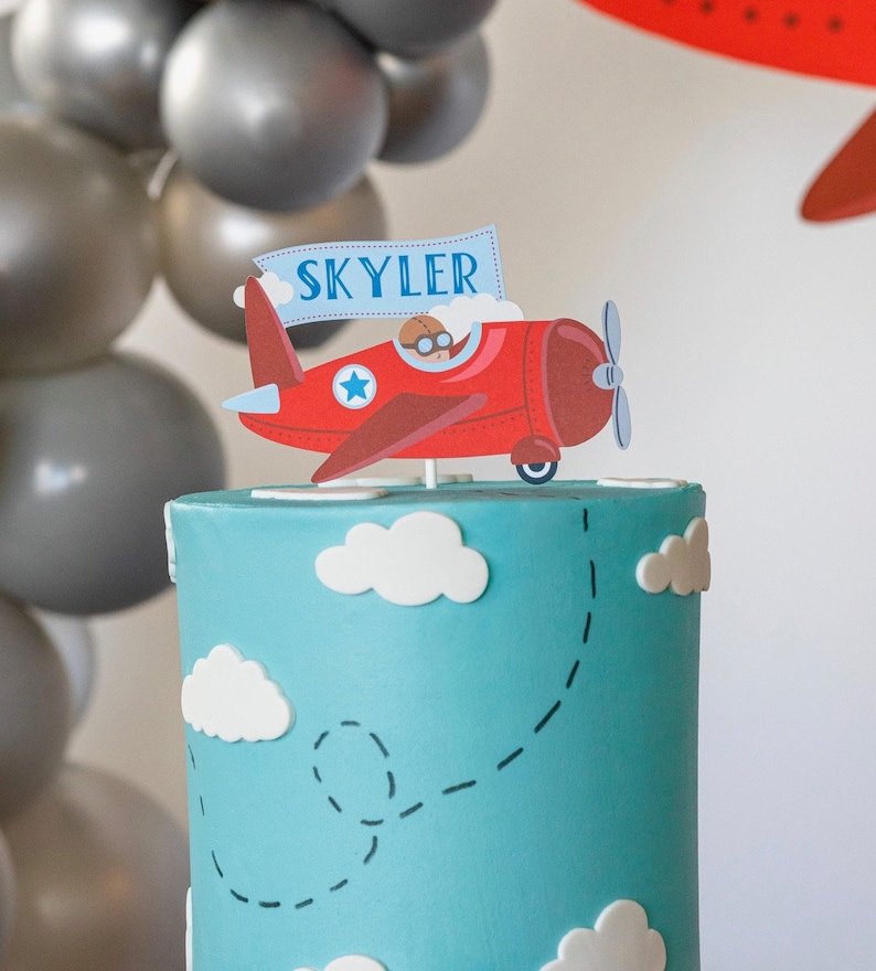 Puede incluir: Una tarta de cumplea&ntilde;os con una base de glaseado azul, decorada con nubes blancas y una trayectoria de vuelo punteada. Un adorno de tarta de avi&oacute;n rojo con el nombre "Skyler" en una pancarta se encuentra encima de la tarta. Globos grises en el fondo.