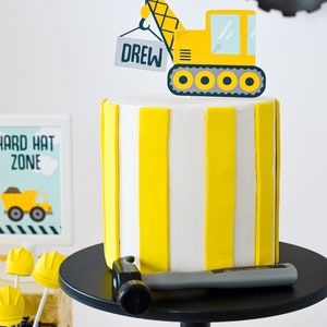 May include: A yellow and white striped cake with a yellow construction crane topper that says "DREW" on the side. A hammer is on a black cake stand.