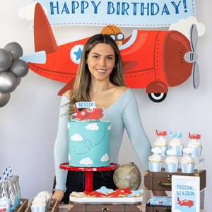 Puede incluir: Decoraci&oacute;n de fiesta de cumplea&ntilde;os con tem&aacute;tica de avi&oacute;n rojo. Una tarta turquesa con nubes blancas y un adorno de avi&oacute;n se encuentra sobre un soporte rojo. Tambi&eacute;n se ven cupcakes, galletas y una pancarta que dice "TIME FLIES".