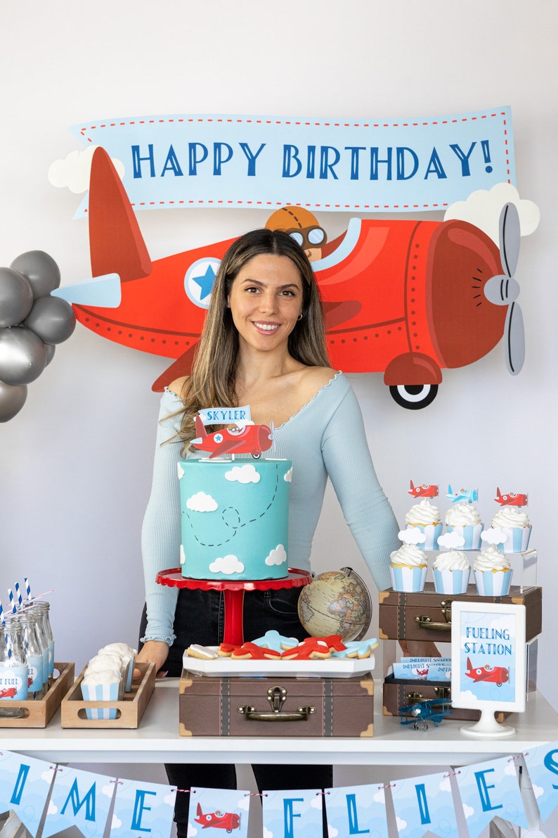 Puede incluir: Decoraci&oacute;n de fiesta de cumplea&ntilde;os con tem&aacute;tica de avi&oacute;n rojo. Una tarta turquesa con nubes blancas y un adorno de avi&oacute;n se encuentra sobre un soporte rojo. Tambi&eacute;n se ven cupcakes, galletas y una pancarta que dice "TIME FLIES".