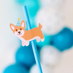 May include: A blue and white striped paper straw with a cartoon corgi wearing a party hat attached to it. The straw is surrounded by blue and white balloons.