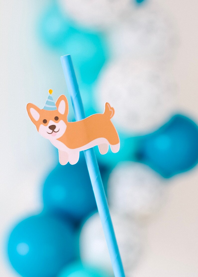 May include: A blue and white striped paper straw with a cartoon corgi wearing a party hat attached to it. The straw is surrounded by blue and white balloons.