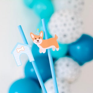 May include: Two blue straws with a dog house and a dog wearing a party hat attached. The straws are in front of a background of blue and white balloons.