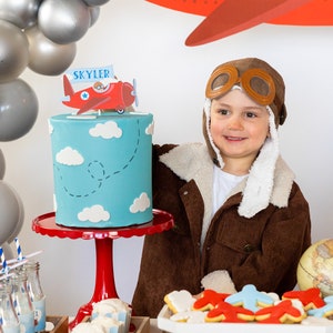Puede incluir: Escena de fiesta de cumplea&ntilde;os con una tarta azul decorada con nubes blancas y un adorno de tarta de avi&oacute;n rojo que dice "Skyler". La tarta est&aacute; sobre un soporte rojo. Otras decoraciones incluyen galletas con forma de avi&oacute;n, globos y un ni&ntilde;o con una chaqueta de aviador marr&oacute;n.