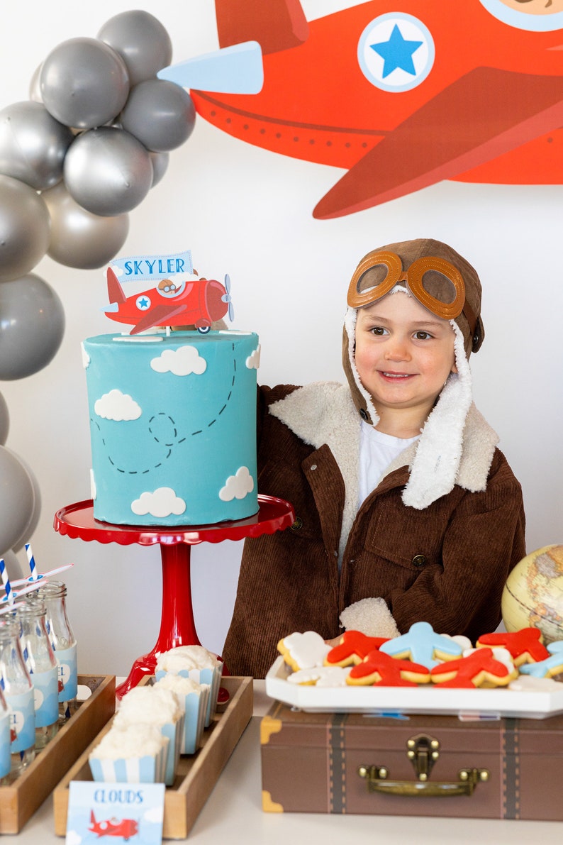 Puede incluir: Escena de fiesta de cumplea&ntilde;os con una tarta azul decorada con nubes blancas y un adorno de tarta de avi&oacute;n rojo que dice "Skyler". La tarta est&aacute; sobre un soporte rojo. Otras decoraciones incluyen galletas con forma de avi&oacute;n, globos y un ni&ntilde;o con una chaqueta de aviador marr&oacute;n.