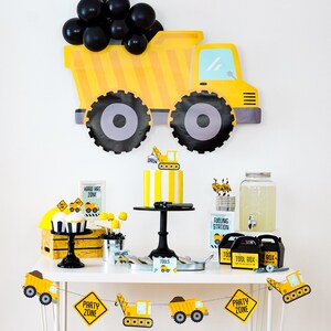 May include: A yellow and black construction truck party decoration with black balloons. A dessert table with a yellow and black striped cake, cupcakes, and other decorations. The table has a sign that says "Hard Hat Zone" and another sign that says "Party Zone".