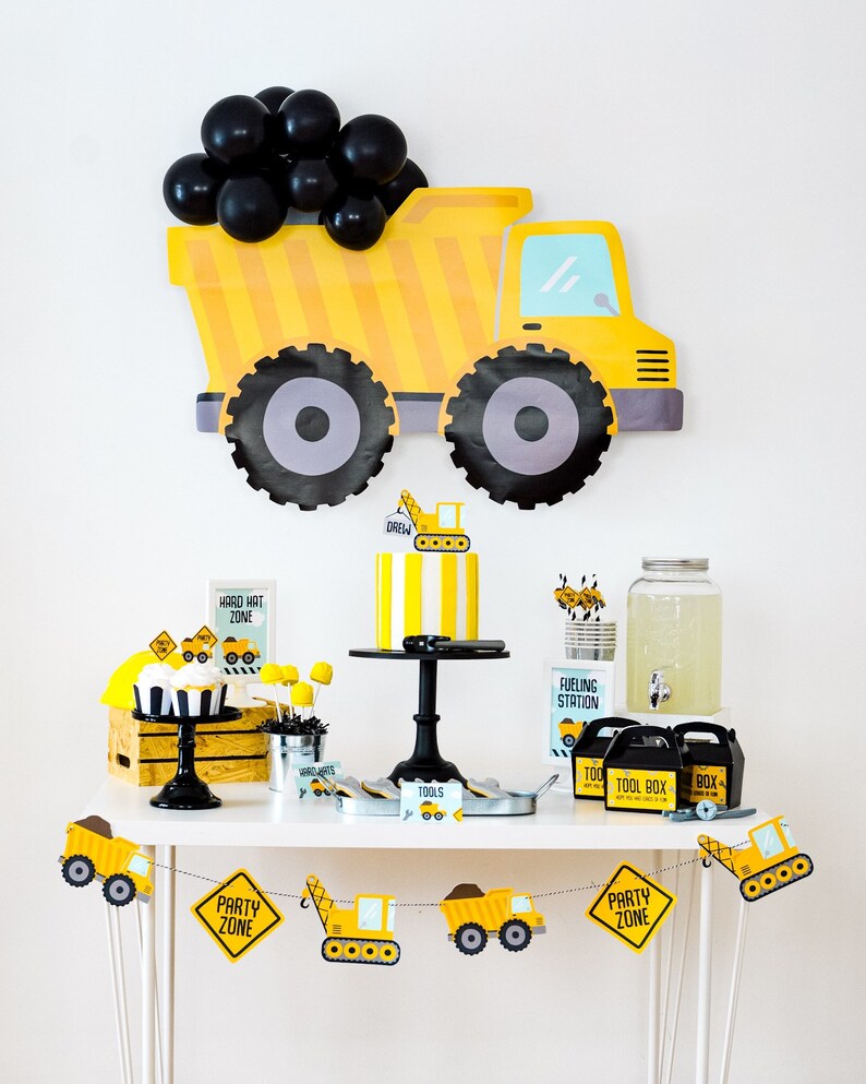 May include: A yellow and black construction truck party decoration with black balloons. A dessert table with a yellow and black striped cake, cupcakes, and other decorations. The table has a sign that says "Hard Hat Zone" and another sign that says "Party Zone".