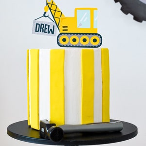 May include: A yellow and white striped cake with a yellow construction vehicle topper. The vehicle is lifting a sign that says "DREW".