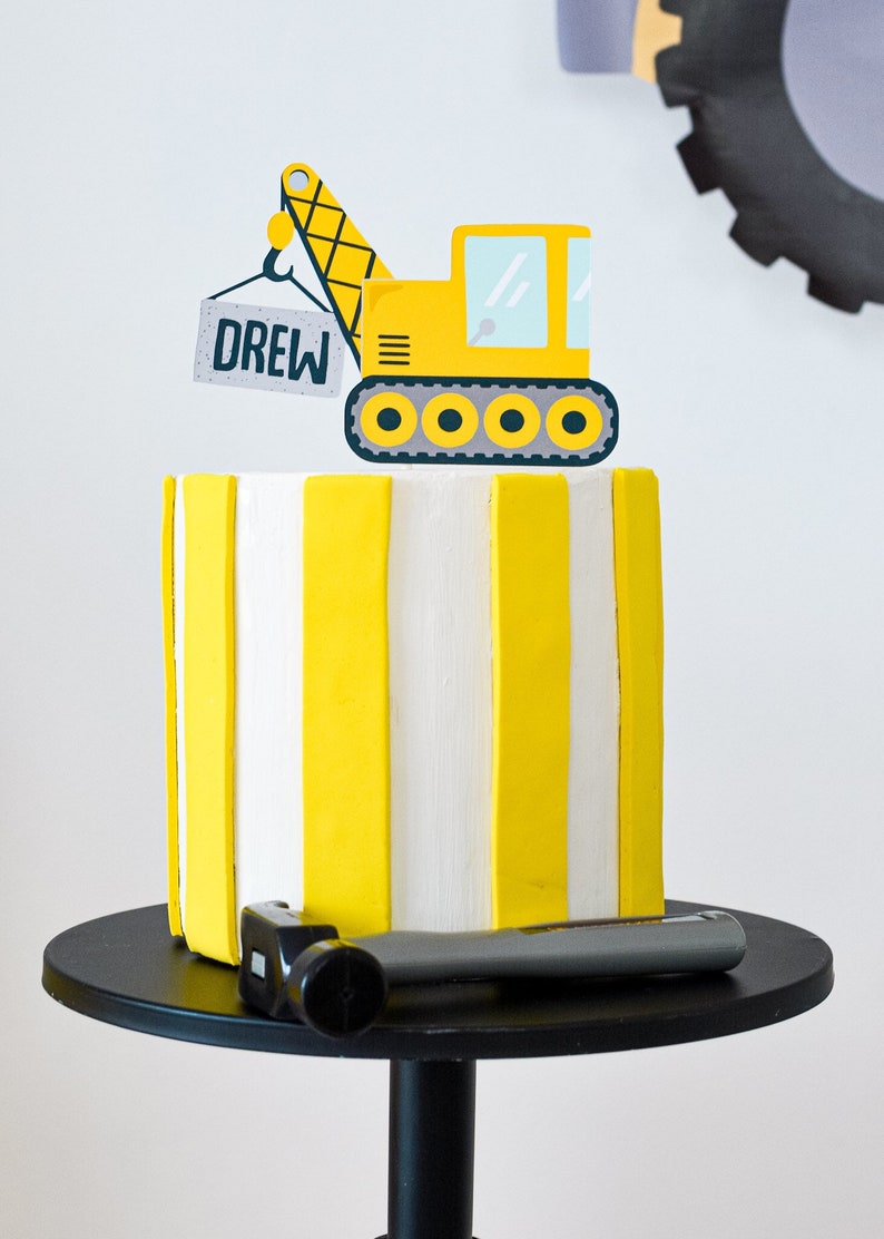 May include: A yellow and white striped cake with a yellow construction vehicle topper. The vehicle is lifting a sign that says "DREW".
