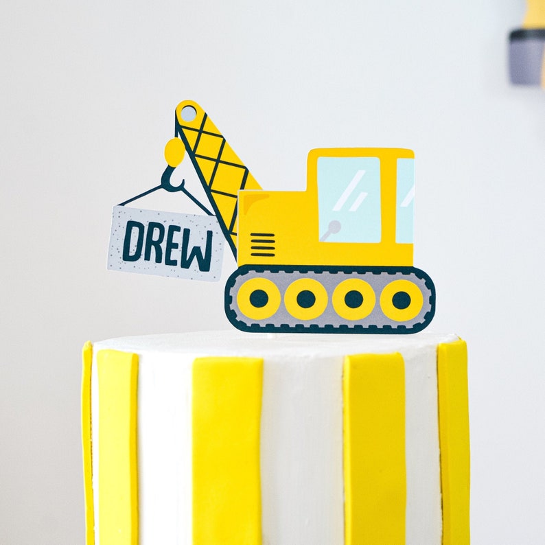 May include: A yellow and black construction vehicle cake topper with the name Drew on a sign. The topper is on top of a white and yellow striped cake.