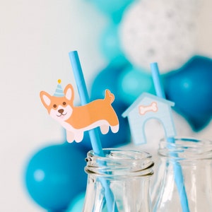 May include: Two clear glass bottles with blue straws. One straw has a paper cutout of a dog wearing a party hat, the other has a paper cutout of a doghouse. The bottles are in front of a background of blue and white balloons.