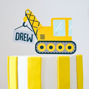 May include: A yellow construction vehicle cake topper with a crane arm and a sign that says "DREW". The vehicle has six wheels and is on a white and yellow striped cake.