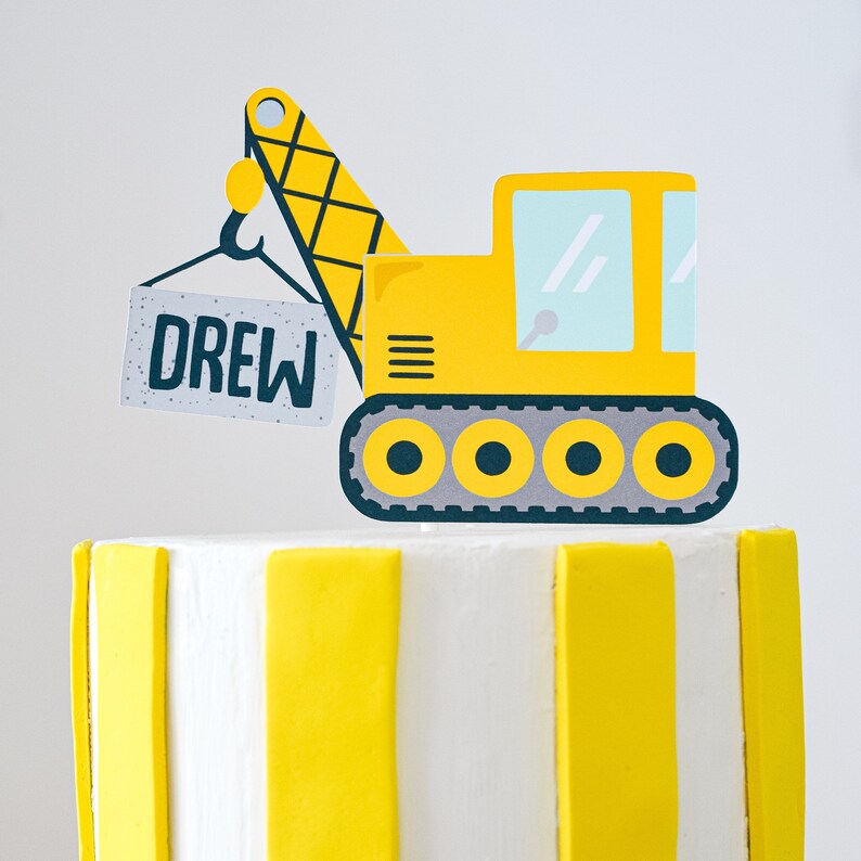 May include: A yellow construction vehicle cake topper with a crane arm and a sign that says "DREW". The vehicle has six wheels and is on a white and yellow striped cake.