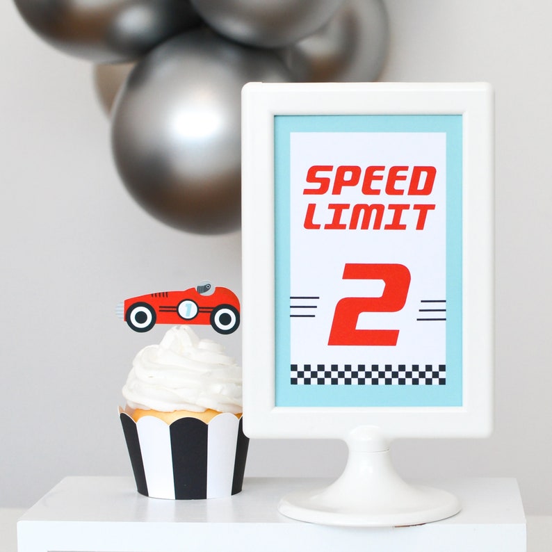 May include: A white cupcake stand with a white frame holding a blue and white sign that says "Speed Limit 2". The sign has a black and white checkered border. A red race car cupcake topper is on top of a black and white striped cupcake.