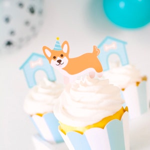 May include: Three cupcakes decorated with white frosting and blue and white striped cupcake liners. The cupcake in the center has a paper cutout of a dog wearing a party hat and a doghouse.