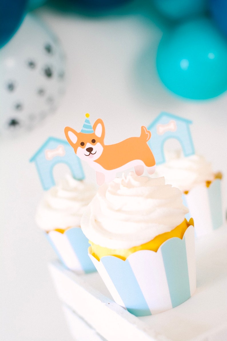 May include: Three cupcakes decorated with white frosting and blue and white striped cupcake liners. The cupcake in the center has a paper cutout of a dog wearing a party hat and a doghouse.