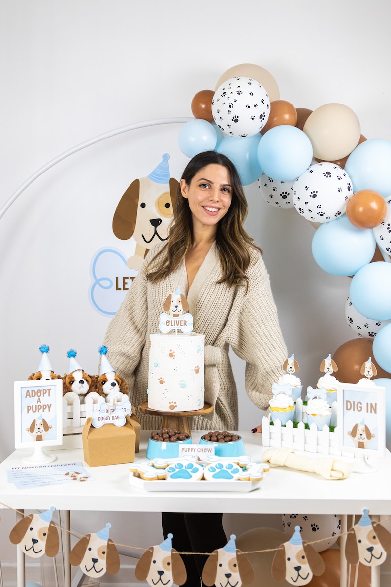 May include: A puppy-themed party setup with a cake, treats, and decorations. The cake is white with paw prints and a topper that says "Oliver." Balloons in blue, brown, and white with paw prints are in the background. There are also signs that say "Adopt a Puppy" and "Dig In."