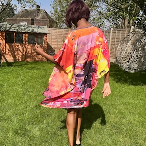 May include: A woman wearing a colorful abstract print kaftan with a flowing design. The kaftan is made of a lightweight fabric and has a relaxed fit. The woman is standing in a grassy area with a wooden shed in the background.