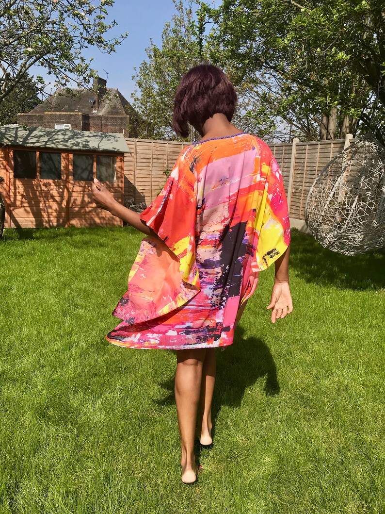 May include: A woman wearing a colorful abstract print kaftan with a flowing design. The kaftan is made of a lightweight fabric and has a relaxed fit. The woman is standing in a grassy area with a wooden shed in the background.