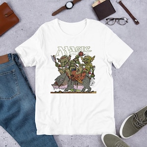 May include: White t-shirt featuring a graphic of goblins riding a large animal, with the text "Magic the Gathering" in green. The goblins are green and brown, and the animal is gray and pink. The shirt is made of a soft material.