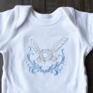 May include: White baby onesie with a blue and white embroidered design of a winged creature with a circular body.
