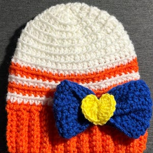 Puede incluir: Gorro de ganchillo hecho a mano en blanco, naranja y azul. El gorro presenta un lazo azul con un corazón amarillo en el centro. El borde y una banda son naranjas, con rayas blancas encima. La parte superior del gorro es blanca.