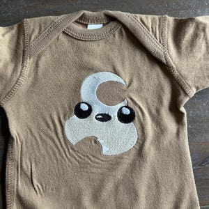 Puede incluir: Un pelele de manga corta marrón con un diseño bordado blanco de un personaje de dibujos animados con ojos grandes y una luna creciente en la cabeza.