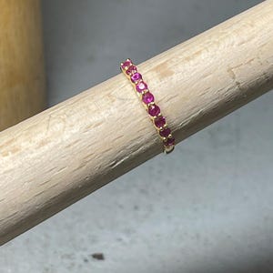 Puede incluir: Un anillo de oro con una fila de pequeñas piedras preciosas redondas de color rosa brillante. El anillo se muestra sobre una clavija de madera, destacando el diseño y el color de la joya.