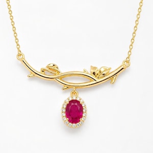 May include: A gold necklace featuring a branch-shaped pendant with small leaves. An oval-cut red gemstone is suspended from the center of the branch, surrounded by a halo of small clear stones. The chain is thin and connects to both ends of the branch.