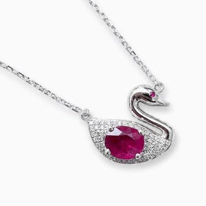 Può includere: Una collana color argento con un ciondolo a forma di cigno. Il corpo del cigno è incastonato con piccole pietre chiare e presenta al centro una grande gemma rosa scuro dal taglio ovale. Una minuscola pietra rosa funge da occhio sulla testa.