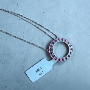 May include: A silver-toned necklace featuring a circular pendant adorned with a ring of small, vibrant pink stones and a border of tiny clear stones. A white rectangular tag is attached to the pendant, displaying the text $850 and #77.