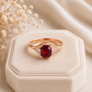 Può includere: Un anello in oro rosa con una pietra preziosa rosso scuro taglio cuscino al centro, affiancata da piccole pietre bianche rotonde incastonate lungo la fascia. L'anello presenta una finitura in metallo lucido e un design simmetrico.