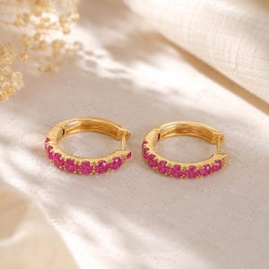 May include: A pair of gold hoop earrings featuring a row of vibrant, round-cut fuchsia pink stones set along the front half of each circular band. The earrings have a hinged closure mechanism and a polished metallic finish on the gold metal.