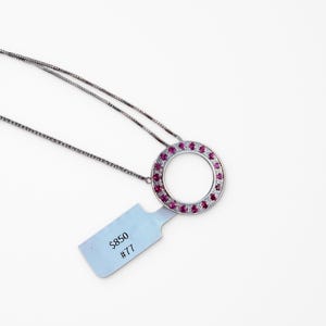 May include: A silver-toned necklace featuring a circular pendant set with a ring of vibrant pink stones and smaller clear stones. A light blue rectangular tag is attached to the pendant, displaying the printed text $850 and #77 in black ink.