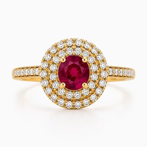 May include: A gold ring featuring a round, deep red centre stone surrounded by a double halo of small, clear, sparkling stones. The band is also set with a row of small, clear stones, creating a cohesive and bright appearance throughout.