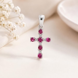 May include: A silver cross pendant featuring five round, vibrant pinkish-red rubies set in a vertical and horizontal arrangement, accented by small, clear, sparkling white stones between each ruby and a polished silver bail at the top.