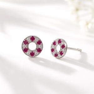 May include: A pair of circular stud earrings featuring a silver-toned metal frame. Each earring is set with alternating round-cut deep pink rubies and small clear white diamonds arranged in a halo pattern around a hollow center post design.
