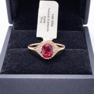 Puede incluir: Un anillo de oro con una gran piedra central de rubí ovalada, rodeada de diamantes más pequeños. El anillo está engastado en una banda de oro y se muestra en una caja de joyería negra. Una etiqueta dice "14K Gold Natural Diamonds Ruby Ring."