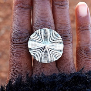 Könnte beinhalten: Ein silberner Ring mit einem runden Design aus konzentrischen Kreisen. Der Ring wird am Mittelfinger einer Hand mit hellbrauner Haut getragen.