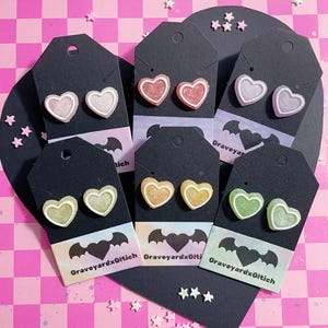 May include: A collection of heart-shaped gummy earrings in various colors, displayed on black tags with a bat graphic and the text "GraveyardxGlitch". The earrings are pink, red, purple, yellow, and green. The background is a pink and white checkered pattern.