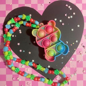 May include: A colorful necklace with a bear-shaped pop-it toy. The necklace features a variety of beads in red, green, blue, and yellow. The pop-it toy has a rainbow gradient color scheme. The background is a black heart shape on a pink checkered surface.